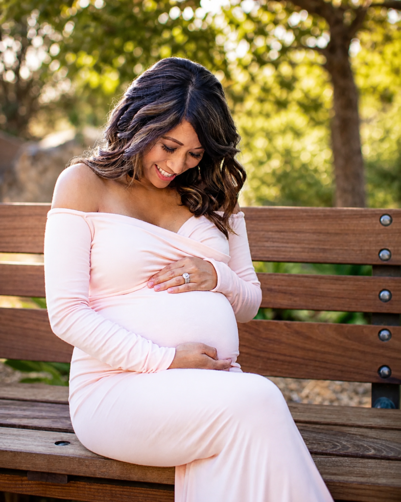 One of my maternity clients wearing a soft pink dress, seated outdoors and cradling her baby bump.
